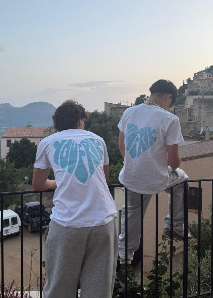 Auf dem Bild sieht man 2 Jungs, die beide das gleiche T-Shirt tragen. Der eine von ihnen sitz auf einem Zaun, während der andere gegen den Zaun lehnt. Sie blicken beide in die Landschaft, in der sich ein altes Bergdorf befindet. Auf den weißen T-Shirts der Jungs steht in Form eines blauen Herzes Youfrik geschrieben. Sie tragen auch beide eine graue Hose