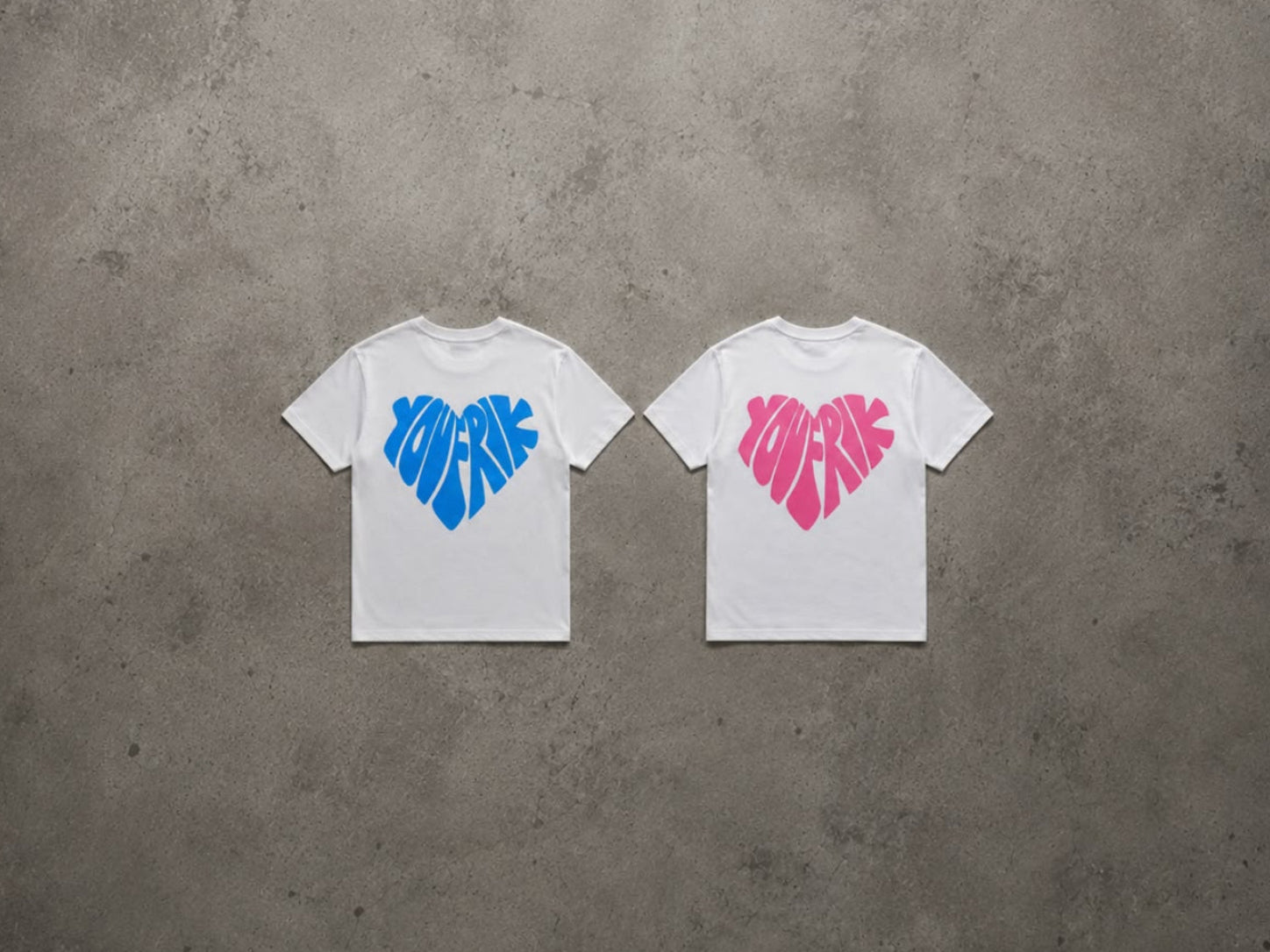 2 T-Shirts liegen auf einem Bettonboden, beide sind weiß, 1 hat einen pinken Schriftzug "Youfrik" in der Form eines Herzes, das andere hat einen blauen Schriftzug "Youfrik" in der Form eines Herzes. 