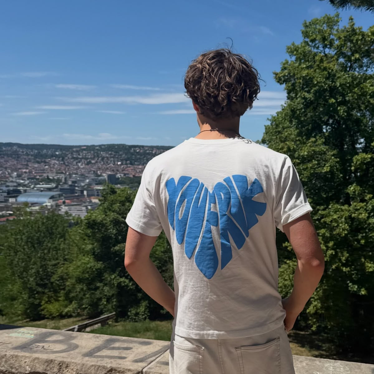 weißes T-Shirt mit blauem herz Backprint wird von einem Mann getragen, der über eine Stadt blickt. 